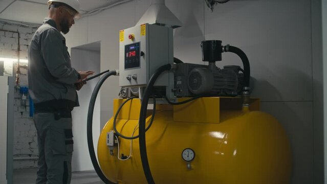 Professional engineer in safety uniform, goggles and protective hard hat adjusts vacuum pump station or air compressor using tablet computer. Male employee works on modern plant or industrial factory.