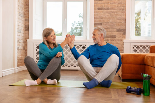 Senior couple sitting on yoga mat in lotus position and giving high five during workout. Two people practicing yoga at home.