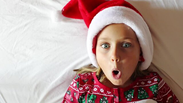 Happy excited funny kid girl in pajamas and Santa on cozy bed with white blanket in Christmas morning or evening. Children are enjoying Christmas holiday. Active child is having fun