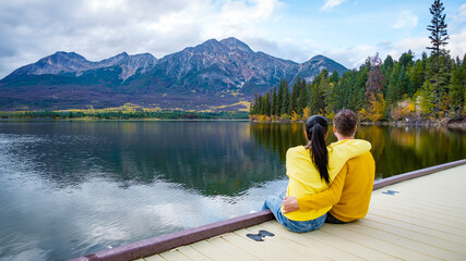 Pyramid Lake Jasper National Park Canadian Rocky Mountains Alberta Canada The Canadian Rockies. Asian women and caucasian men sitting on a wooden pier arms together at Pyramid Lake Autumn fall season - obrazy, fototapety, plakaty