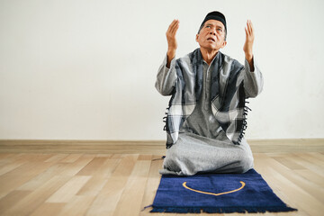 Elderly Asian muslim man sitting on prayer mat, raising hands and looking up praying to God