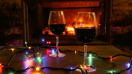 Two red wine glasses and lights decoration on burning fireplace background. Wine by the fireplace.