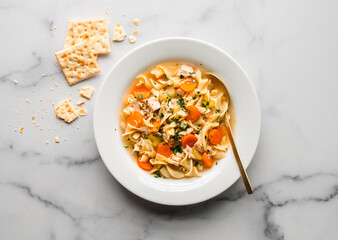 Top view bowl of chicken noodle soup on white marble backdrop.