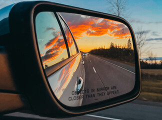 Sunset reflected in side mirror of vehicle on tree lined road.