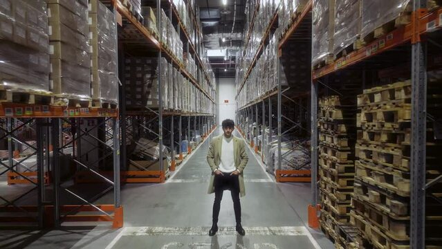 Handsome Smiling Boss with Arms in Pockets, Stands in Retail Warehouse Full of Shelves Packed with Cardboard Boxes. Professional Leader in Logistics, Delivery, and Distribution Center