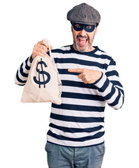 Middle age handsome man wearing burglar mask holding money bag smiling happy pointing with hand and finger