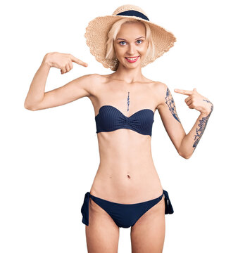 Young blonde woman with tattoo wearing bikini and summer hat looking confident with smile on face, pointing oneself with fingers proud and happy.