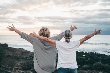 Back view of mature and senior women standing face to the sea with outstretched arms looking at horizon over water, two bonding people stay together enjoying freedom and vacation or retirement