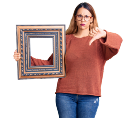 Beautiful young woman holding empty frame with angry face, negative sign showing dislike with thumbs down, rejection concept