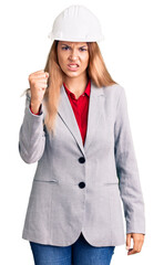 Beautiful young woman wearing architect hardhat angry and mad raising fist frustrated and furious while shouting with anger. rage and aggressive concept.