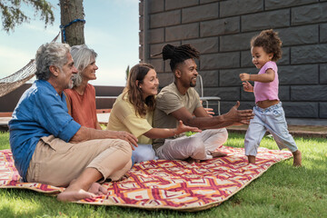 A multiracial family enjoys a picnic day as a toddler joyfully runs towards her parents and...