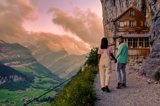 A couple of men and women hiking to Berggasthaus Aescher in den Appenzeller Alpen at sunset, a restaurant under a cliff at the mountain Ebenalp in Switzerland, Appenzell