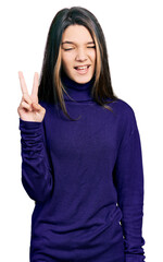 Young brunette girl with long hair wearing turtleneck sweater smiling with happy face winking at the camera doing victory sign. number two.