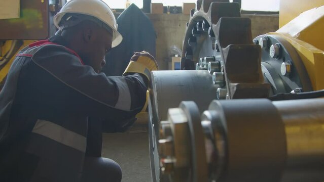 Side shot of African American factory worker screwing up nuts on large cogwheel on tractor framework using wrench at heavy machinery production plant