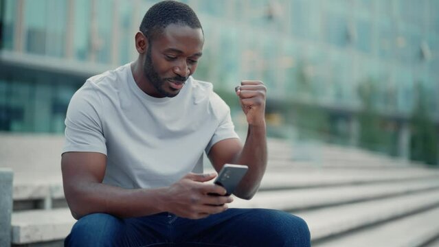 Overjoyed amazed african american man showing win victory hands gesture looking at smartphone screen. Smiling guy winner celebrates success, achieving goal. Body language, sincere emotions concept.