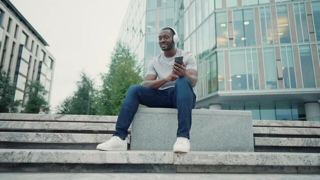 African american man music lover in headphones enjoying listening music on smartphone sitting on city street outdoors. Positive smiling guy melomaniac rhythmically nodding head relaxing moving body.