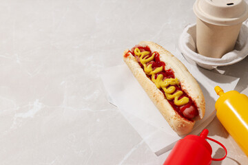 Hot dog, sauces in bottles and paper cup on light background, space for text
