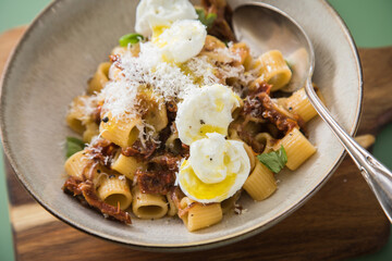 Italienische mini Burrata Käse Kugeln mit Mezzi Rigatoni Pasta, getrockneten Tomaten, Parmesan, Olivenöl, Basilikum, Knoblauch und Chili Flocken in Schale mit Löffel, Holzbrett und grün Hintergrund