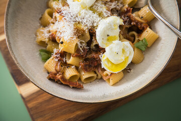 Italienische mini Burrata Käse Kugeln mit Mezzi Rigatoni Pasta, getrockneten Tomaten, Parmesan, Olivenöl, Basilikum, Knoblauch und Chili Flocken in Schale mit Löffel, Holzbrett und grün Hintergrund