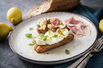 Italienische Burrata Käse Crostini aus Ciabatta Brot,konfierter Knoblauch, Prosciutto Schinken, rote Zwiebel, Basilikum, Honig, Olivenöl, Zitronen mit Teller, Besteck, Serviette blau, dunkel grau Stei