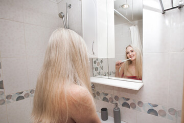 Smiling sexy blonde woman in a red towel brushes her teeth with an electric toothbrush, in the...