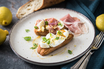 Italienische Burrata Käse Crostini aus Ciabatta Brot,konfierter Knoblauch, Prosciutto Schinken, rote Zwiebel, Basilikum, Honig, Olivenöl, Zitronen mit Teller, Besteck, Serviette blau, dunkel grau Stei