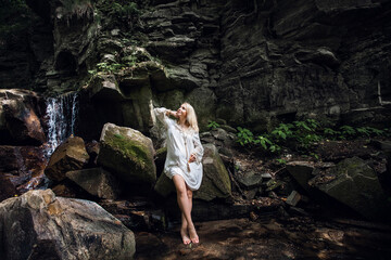 A smiling happy pregnant girl in a light vintage dress, with blond short hair, stands against the backdrop of picturesque rocks near a waterfall in the forest.