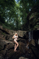 A smiling happy pregnant girl in light underwear, with blond short hair, stands against the backdrop of picturesque rocks near a waterfall in the forest.