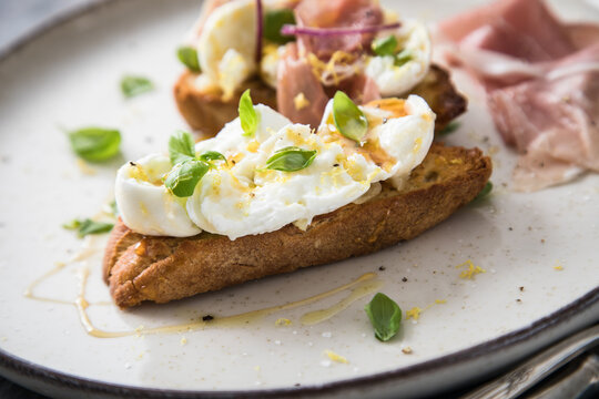Italienische Burrata K&auml;se Crostini aus Ciabatta Brot,konfierter Knoblauch, Prosciutto Schinken, rote Zwiebel, Basilikum, Honig, Oliven&ouml;l, Zitronen mit Teller, Besteck, Serviette blau, dunkel grau Stei