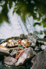 A slender sweet girl with blond hair, tanned skin, dressed in a green stylish swimsuit and with a hat, sits on stones against the backdrop of a picturesque waterfall and rests in the summer.