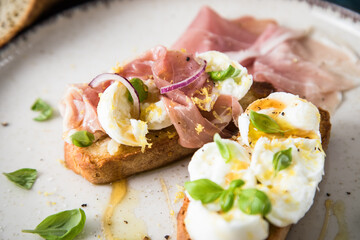 Italienische Burrata Käse Crostini aus Ciabatta Brot,konfierter Knoblauch, Prosciutto Schinken, rote Zwiebel, Basilikum, Honig, Olivenöl, Zitronen mit Teller, Besteck, Serviette blau, dunkel grau Stei