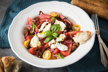 Italienischer Burrata Käse Salat mit bunten roten und gelben Tomaten, Basilikum, Ciabatta Brot Olivenöl und Balsamico Dressing mit tiefer Teller, Besteck Serviette blau, dunkel grau Stein Hintergrund