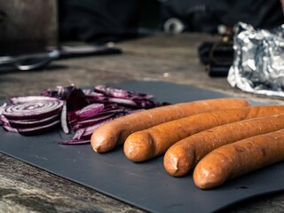 Cutting board on an outdoor wooden camping table with cut red onion, mushroom, garlic, black hunting knife, tin foil, sausages hot-dogs