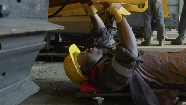 Side shot of African American workman rolling on wheels under tractor frame and tensioning bolts or nuts with wrench at manufacturing plant