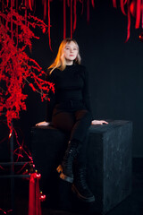 Obraz na płótnie Canvas A beautiful young blonde girl in New Year's black clothes in an atmospheric photo studio with a stylish festive interior in red colors.