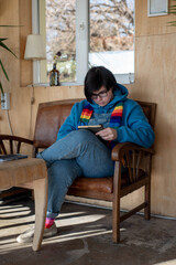 Concentrated androgynous teenager is lying comfortably in an armchair with a book