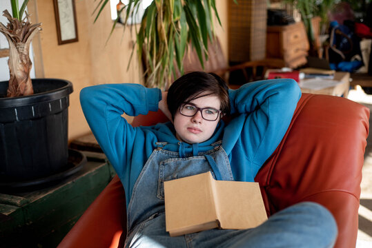 Pensive androgynous teenager is lying comfortably in an armchair with a book