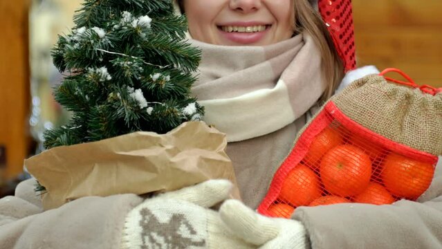 A beautiful young blonde woman at a Christmas fair. The woman is holding a small Christmas tree and brightly coloured tangerines. Merry Christmas.