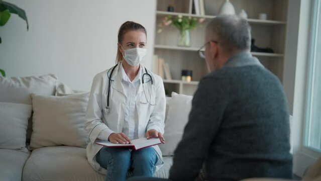 Woman doctor geriatric in medical mask holding notepad consulting senior man in clinic. Medical practitioner ask questions, filling patient disease history form, provide help, prescribe treatment.