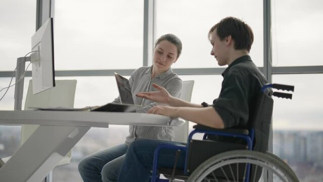 Young male office worker in a wheelchair talking to female colleague while showing her ideas using laptop in co-working space. Disability and handicap concept. Office modern people at work.