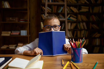 Cute little elementary school boy student hiding behind copybook