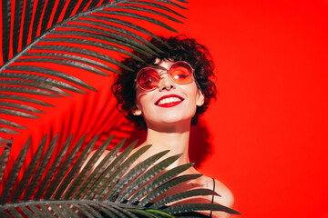 Fashion beauty portrait of young smiling brunette woman with evening stylish  makeup. Sexy model with curly short hair posing in studio. With red bright lips. Tropical palm leaf. In sunglasses