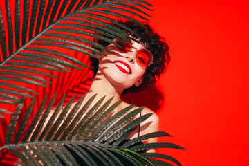 Fashion beauty portrait of young smiling brunette woman with evening stylish  makeup. Sexy model with curly short hair posing in studio. With red bright lips. Tropical palm leaf. In sunglasses