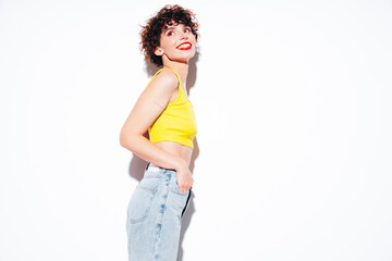 Young beautiful smiling female in trendy summer yellow tank top. Carefree woman posing near white wall in studio with curly hairstyle. Positive model having fun, going crazy. Cheerful and happy