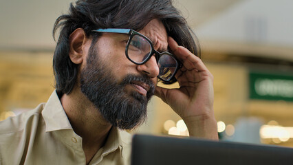 Indian bearded stressed man in glasses working at office with laptop Arabian businessman sad with business problem failure head pain suffering from chronic severe headache thinking trouble solution