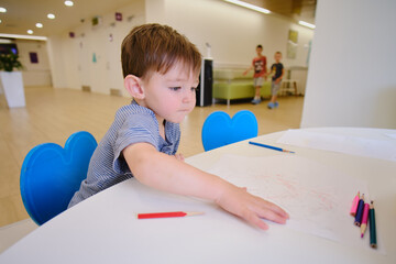 The child is learning and developing their creativity by drawing with a pencil on a piece of paper in the hall of the medical center while waiting for their turn to see the doctor. Kid aged two years