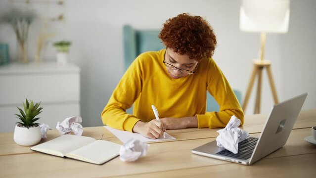 Young woman journalist writer in glasses writing article book on paper shits sitting at table near laptop. Crumpled papers on table around tired girl. Writing novel, letter, creating process concept.