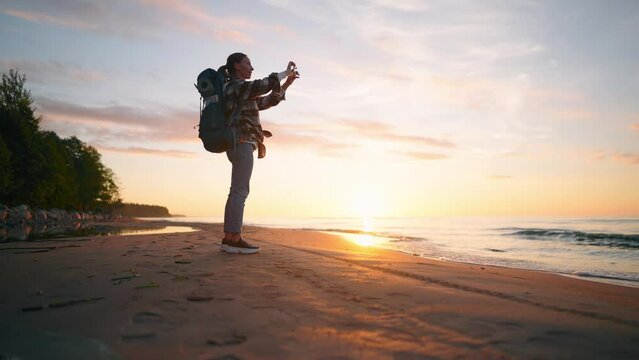 Woman tourist takes photo of sea sunset. Female hiker camper with touristic backpack recording video of nature on smartphone enjoying vacations. Travel tourism trip hiking camping wanderlust concept.