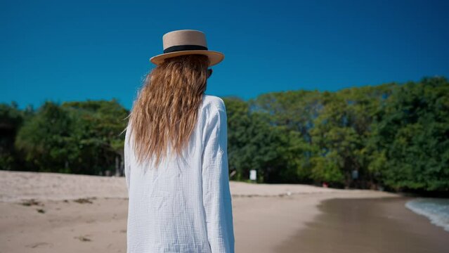 Blonde woman in white airy tunic, straw hat walking along sandy beach looking at ocean enjoy summer tropical vacations. Tourist enjoying resting by sea. Tourism travel journey trip concept.