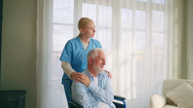 Old man in wheelchair talks with nurse. Senior paralyzed male thanks social worker woman for help support at home. Caregiver holds hand on patient shoulder stands near. Legs paralysis, special needs.